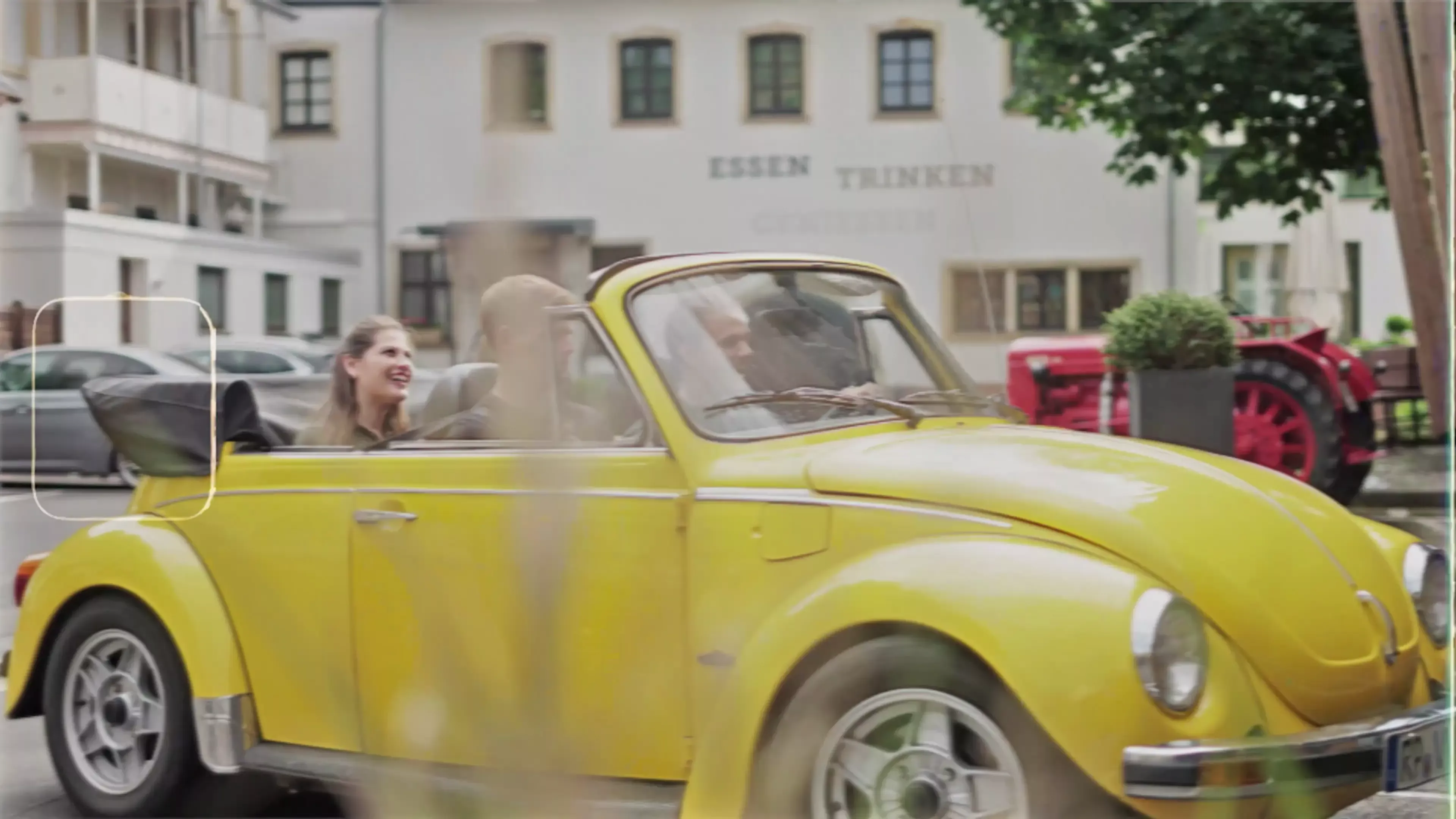 Eine lächelnde Frau und ein Mann fahren mit einem leuchtend gelben Oldtimer-Cabrio an einem Gebäude mit der Aufschrift "ESSEN TRINKEN" an der Wand vorbei. Die Straße ist begrünt und ein rotes Fahrzeug ist im Hintergrund geparkt.
