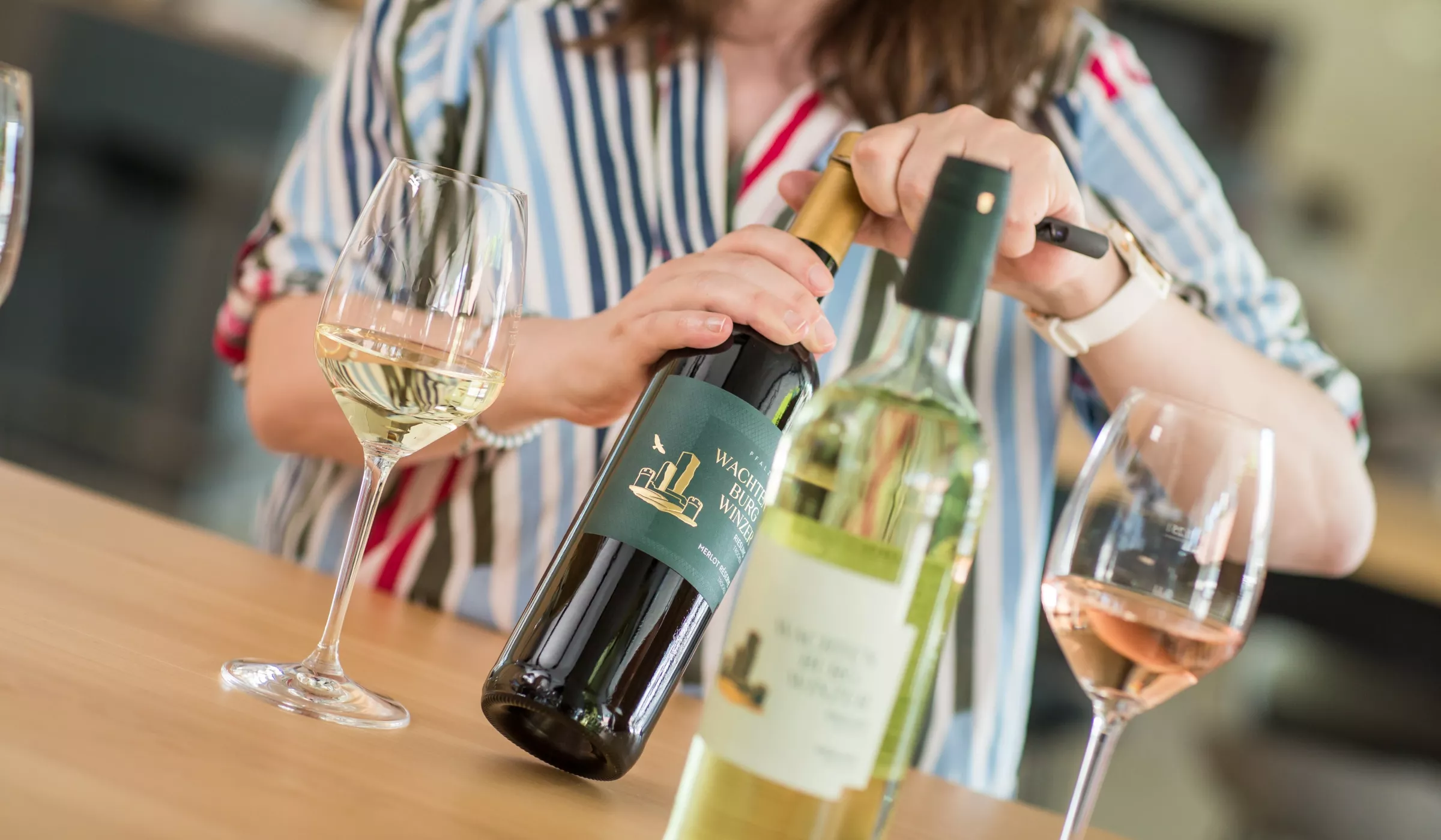 Eine Person in einem gestreiften Hemd öffnet an einem Tisch eine Weinflasche, daneben stehen zwei Weingläser und eine weitere Flasche.