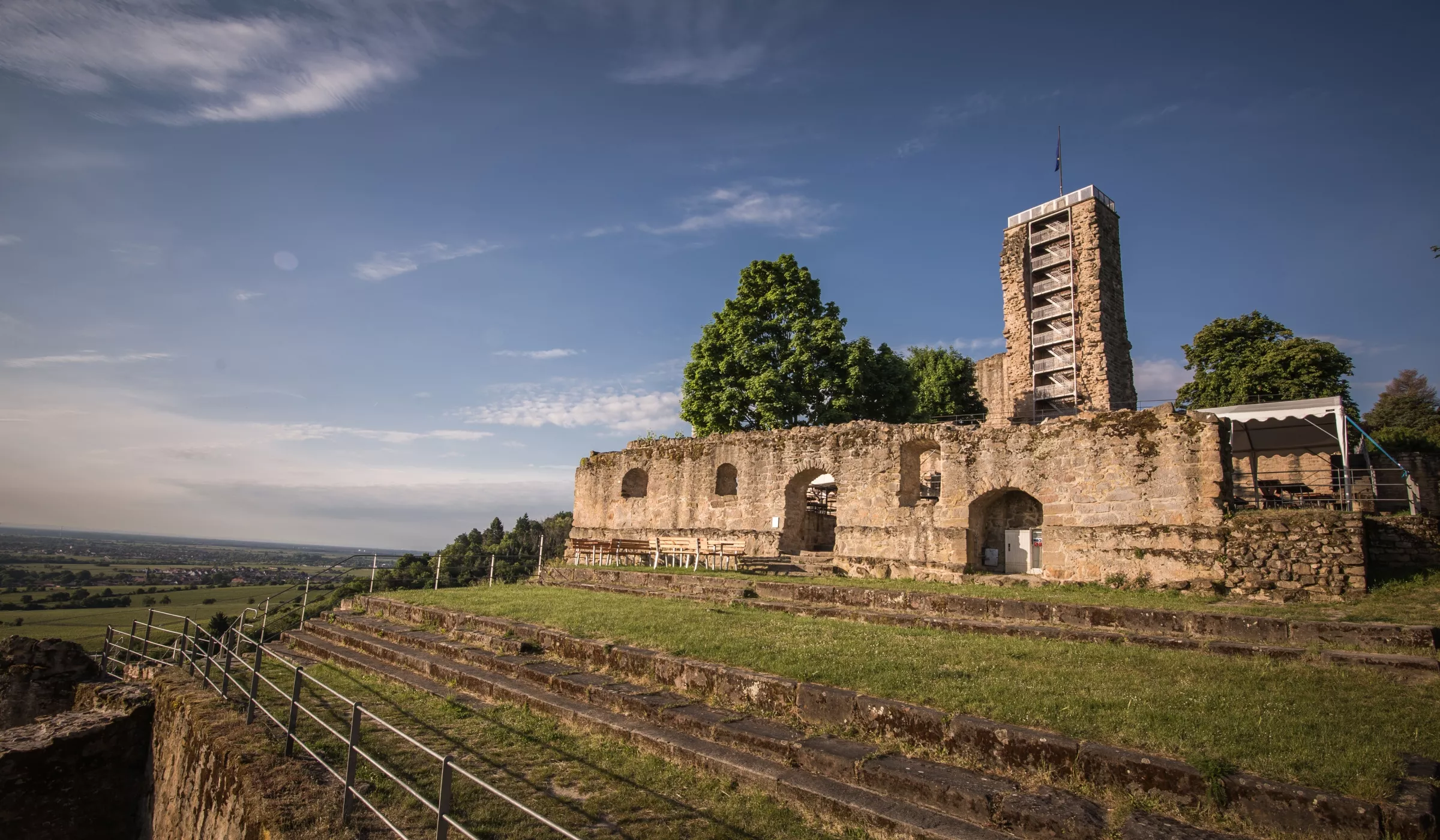 Eine alte Steinruine mit gewölbten Toren und einem hohen modernen Aussichtsturm dahinter, an einem grasbewachsenen Hang gelegen, mit Blick auf eine weite, grüne Landschaft unter einem teilweise bewölkten Himmel.