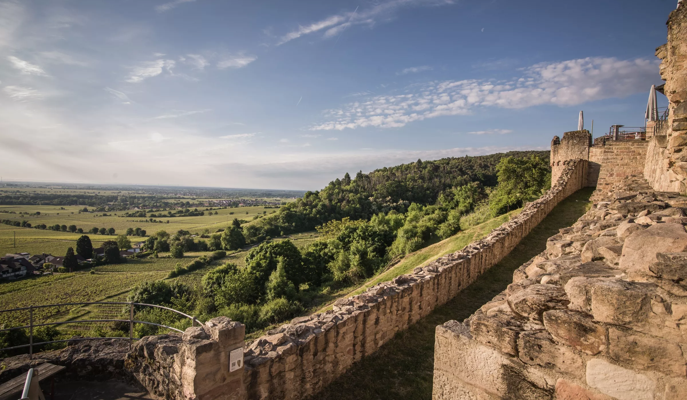 Die steinernen Burgmauern erstrecken sich entlang einer Hügelkuppe und bieten einen Blick auf ein üppig grünes Tal und ferne Felder unter einem blauen Himmel mit vereinzelten Wolken. Bäume und Vegetation umgeben die historische Stätte.