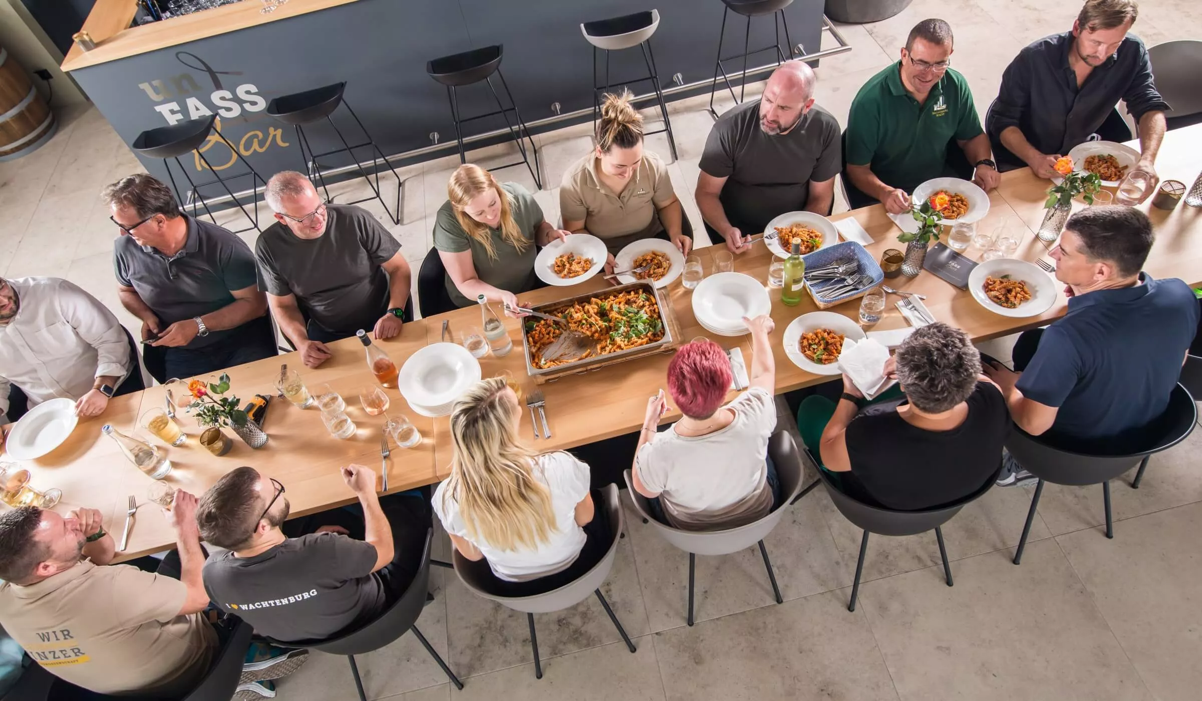 Eine Gruppe von Menschen sitzt an einem großen Holztisch und isst gemeinsam, jeder hat einen Teller mit Essen. Auf dem Tisch stehen Teller, Gläser und ein großes Tablett mit Nudeln. Die Szene wirkt zwanglos und gesellig, in einem modernen, hellen Raum.