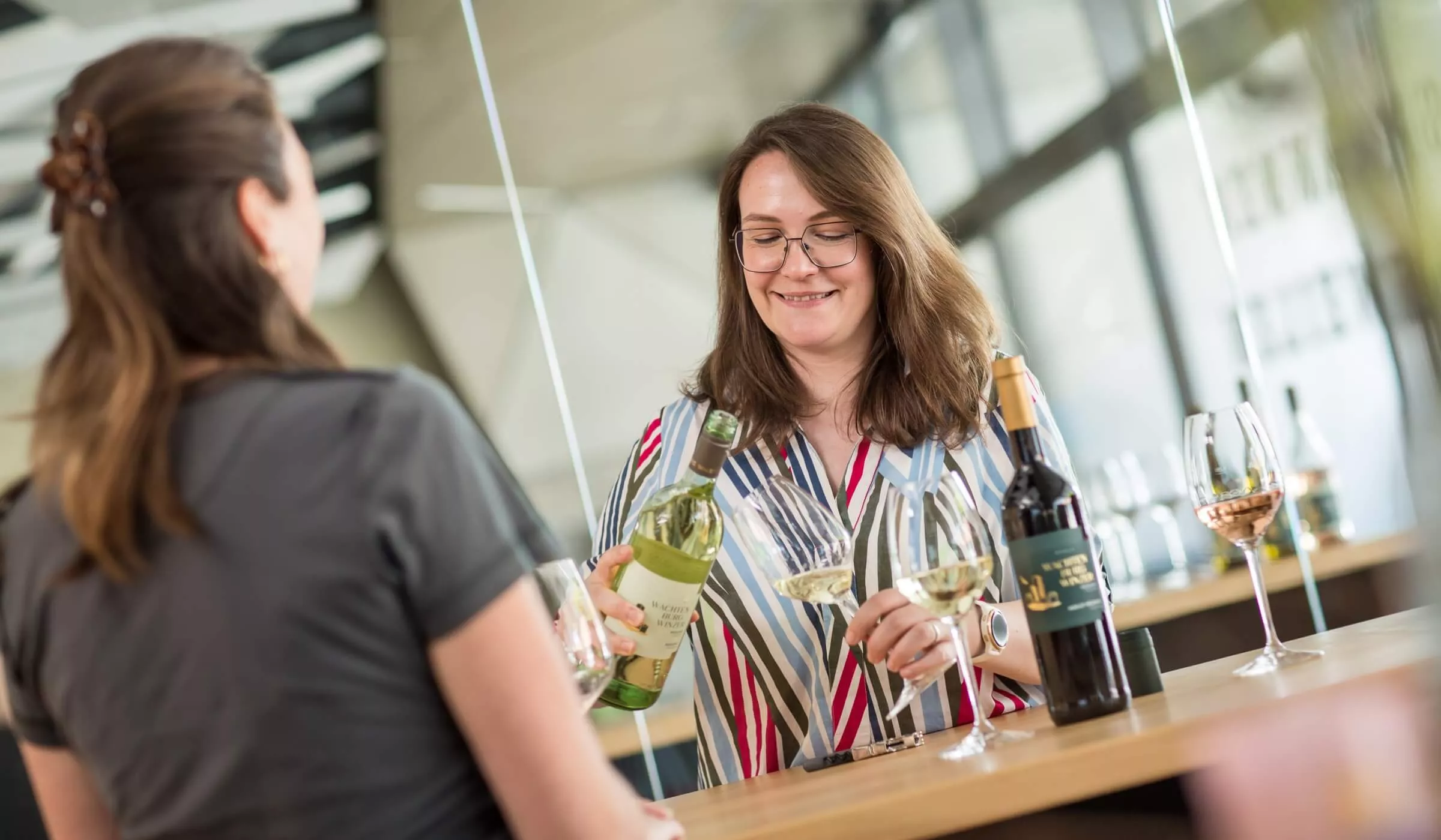 Eine Frau gießt Weißwein in ein Glas für eine andere Person in einer modernen Weinverkostungsbar ein, auf deren Tresen Flaschen und gefüllte Gläser stehen.