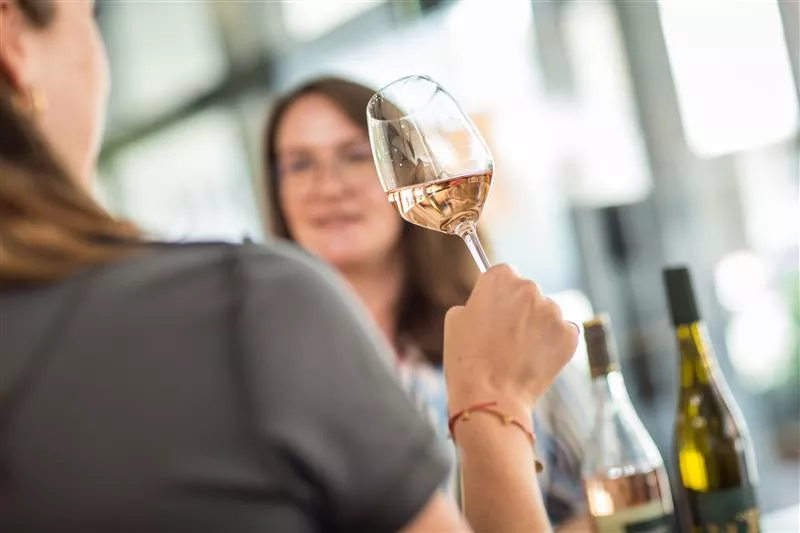 Eine Person, die ein Glas Roséwein in der Hand hält, unterhält sich mit einer anderen Person in einem hellen Raum, in dem Weinflaschen auf dem Tisch stehen.