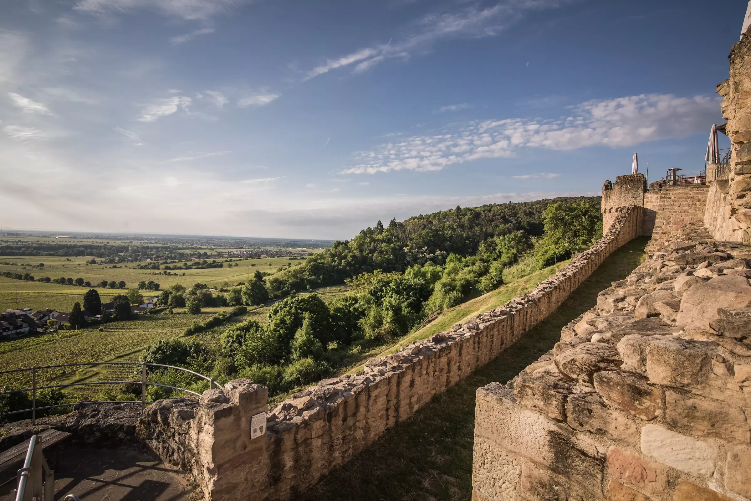 Eine steinerne Burgmauer schlängelt sich an einer Hügelkuppe entlang und bietet einen Blick auf grüne Felder, Bäume und entfernte Ackerflächen unter einem teilweise bewölkten blauen Himmel.