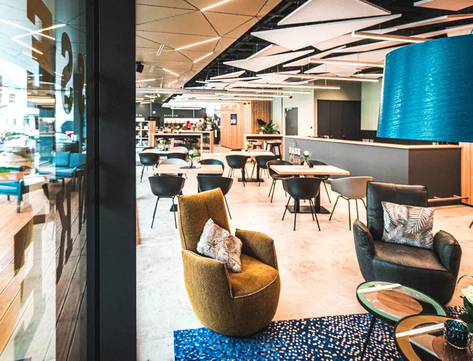 Moderne, stilvolle Bar mit gemütlichen Sesseln, runden Tischen, schwarzen Stühlen und geometrischen Deckenplatten. Ein blauer Lampenschirm und ein Teppich bringen Farbe ins Spiel, während große Fenster natürliches Licht hereinlassen. Pflanzen und dekorative Kissen schaffen eine einladende Atmosphäre.