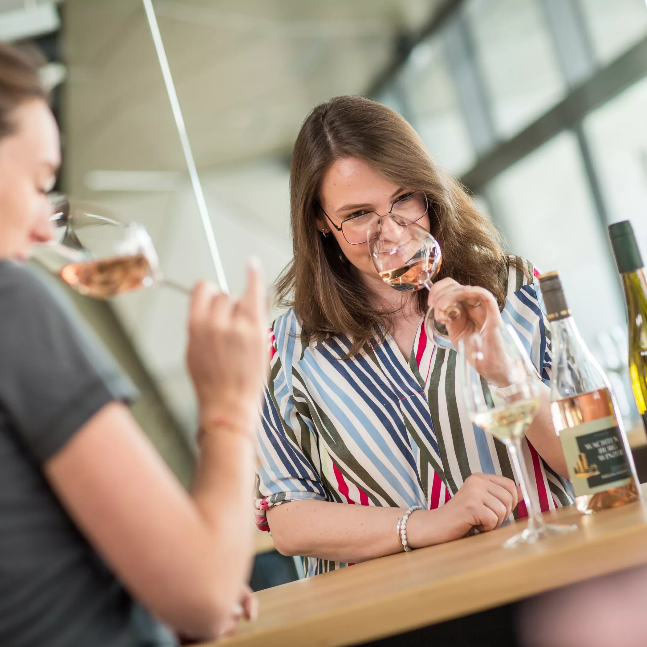 Zwei Frauen stehen an einer Bar, lächeln und riechen an Gläsern mit Weiß- und Roséwein. Auf dem Tresen stehen Weinflaschen. Der Hintergrund ist hell und modern und deutet auf eine Weinprobe oder eine gesellschaftliche Veranstaltung hin.