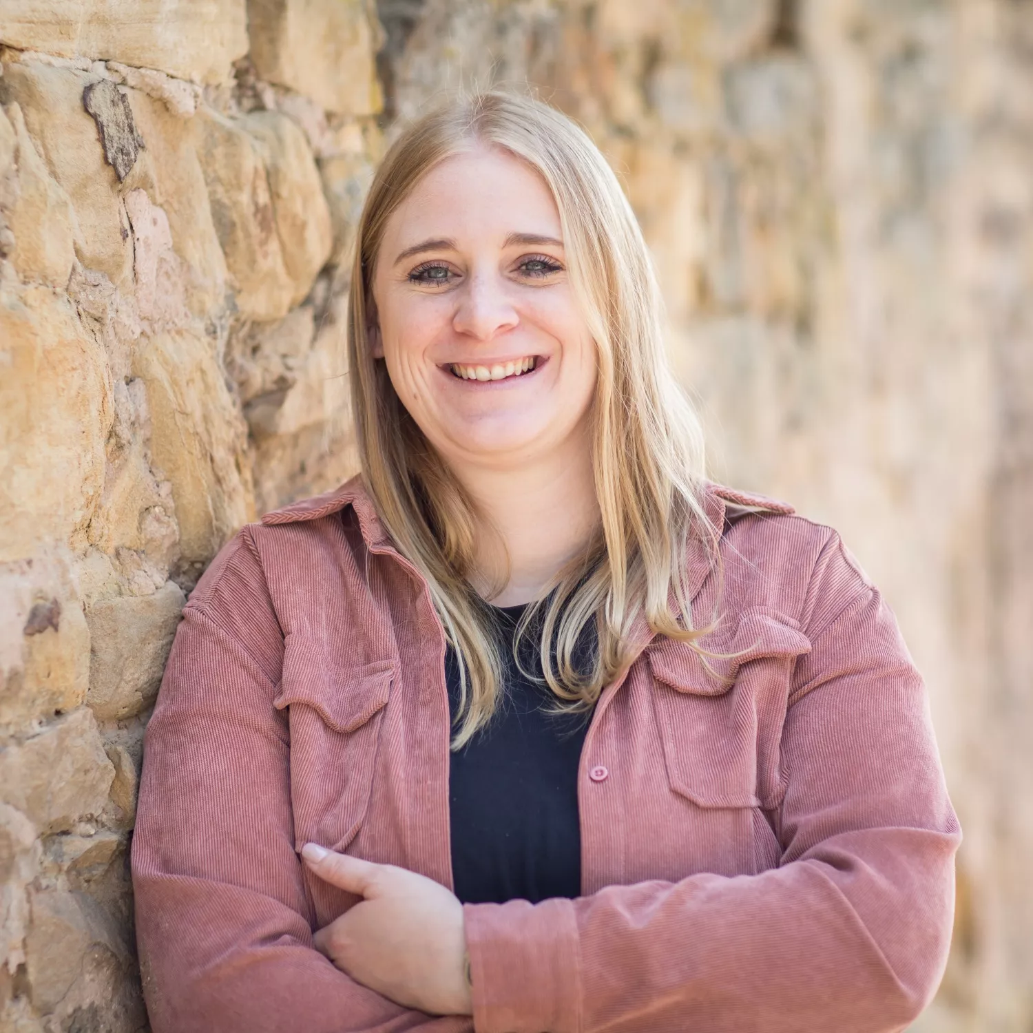 Eine lächelnde Frau mit langen blonden Haaren, die eine lilafarbene Jacke über einem schwarzen Oberteil trägt, steht mit verschränkten Armen vor einer Steinmauer im Tageslicht.