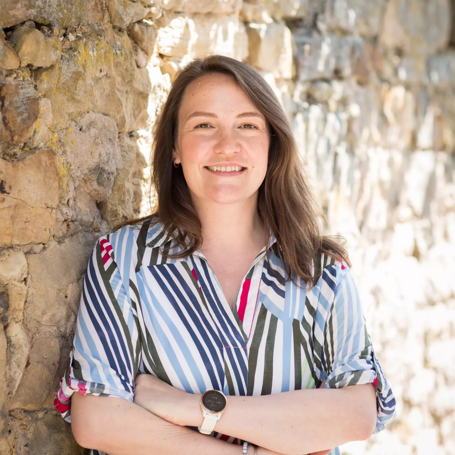 Eine Frau mit langen braunen Haaren, einem gestreiften Hemd und einer Uhr steht mit verschränkten Armen vor einer sonnenbeschienenen Steinmauer und lächelt.