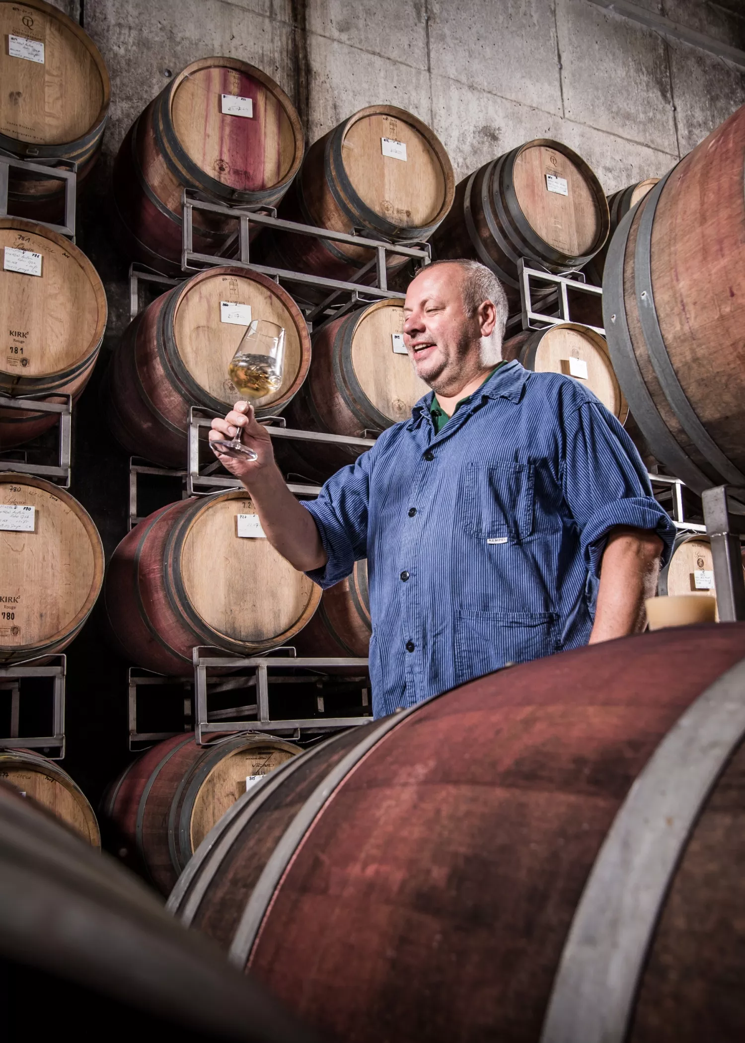 Ein Mann in einem blauen Hemd prüft ein Glas Weißwein, während er in einem mit gestapelten Holzfässern gefüllten Keller steht.