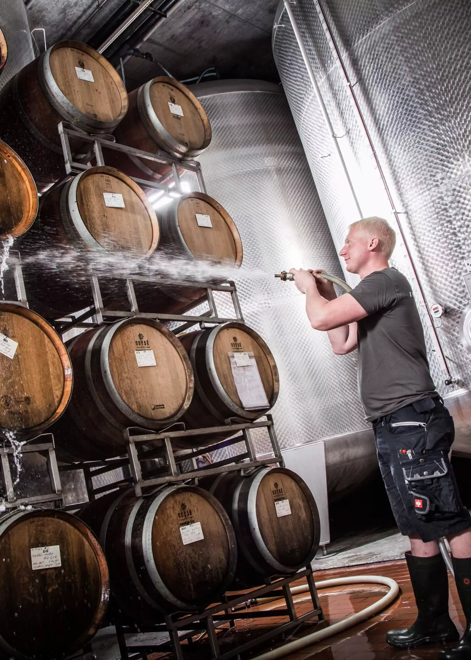 Eine Person in Arbeitskleidung sprüht Wasser auf gestapelte Holzfässer in einer Weinkellerei oder Brauerei, mit großen Metalltanks im Hintergrund.